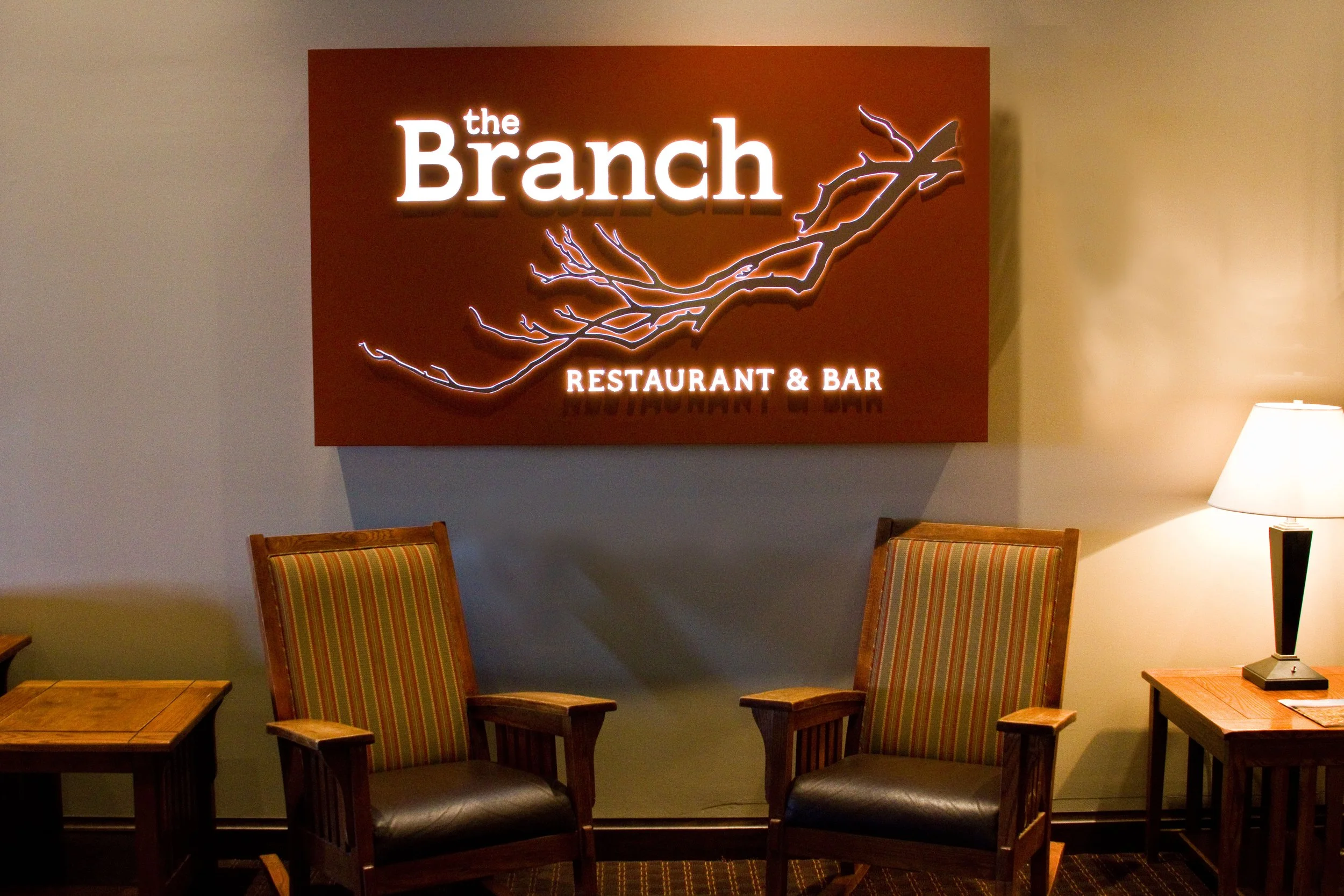 A sign that reads 'the Branch' with a branch illustration and 'Restaurant & Bar' underneath, surrounded by four wooden chairs with striped cushions, a small table, and a table lamp in an indoor setting.