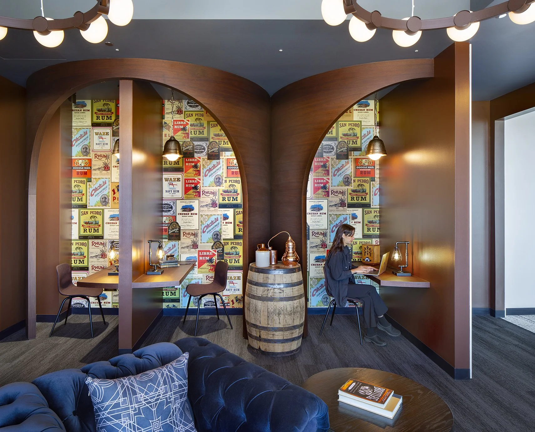Interior of a modern room with dark wood and the walls decorated with vintage-style posters, featuring a woman sitting at a desk working on a laptop, two chairs and a wooden barrel in the space, bar-style lighting, and a plush blue sofa with a geomet