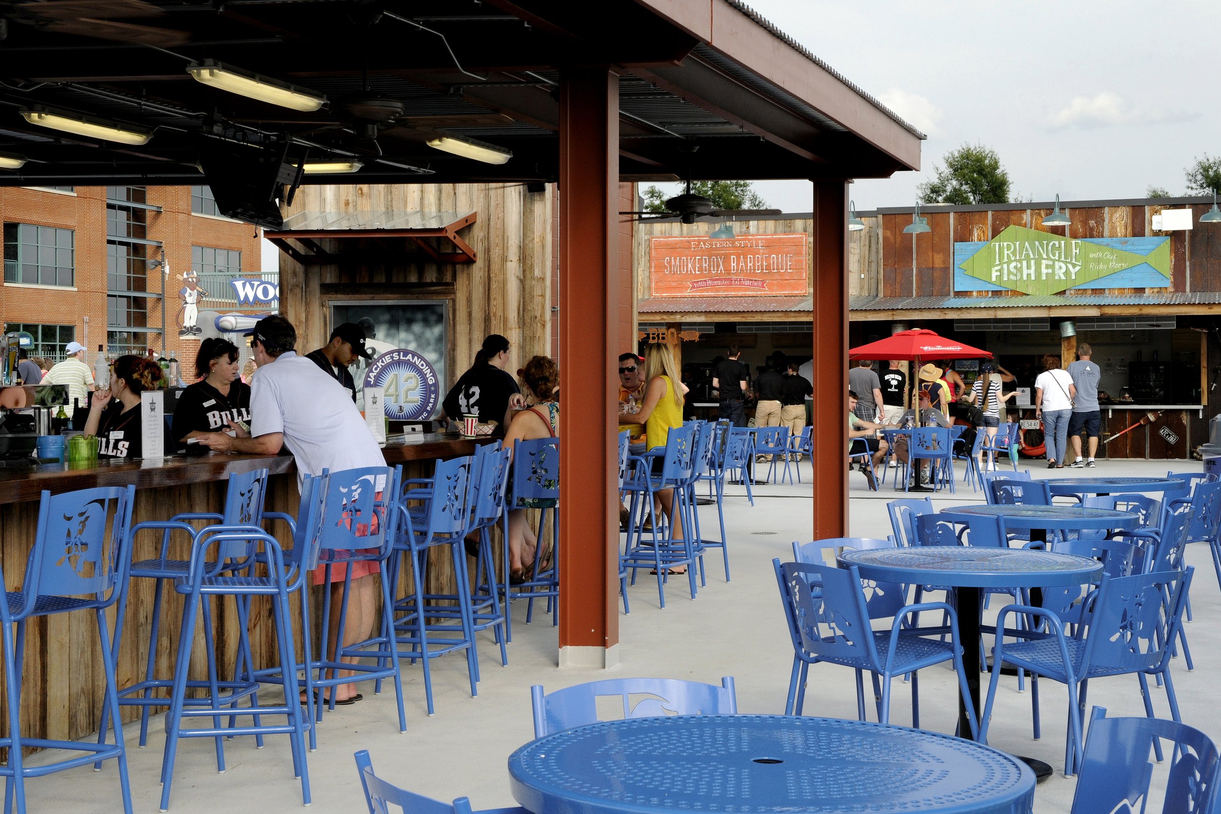 Outdoor dining area with blue chairs, people ordering food at a counter, and signs for barbecue and fish fry.