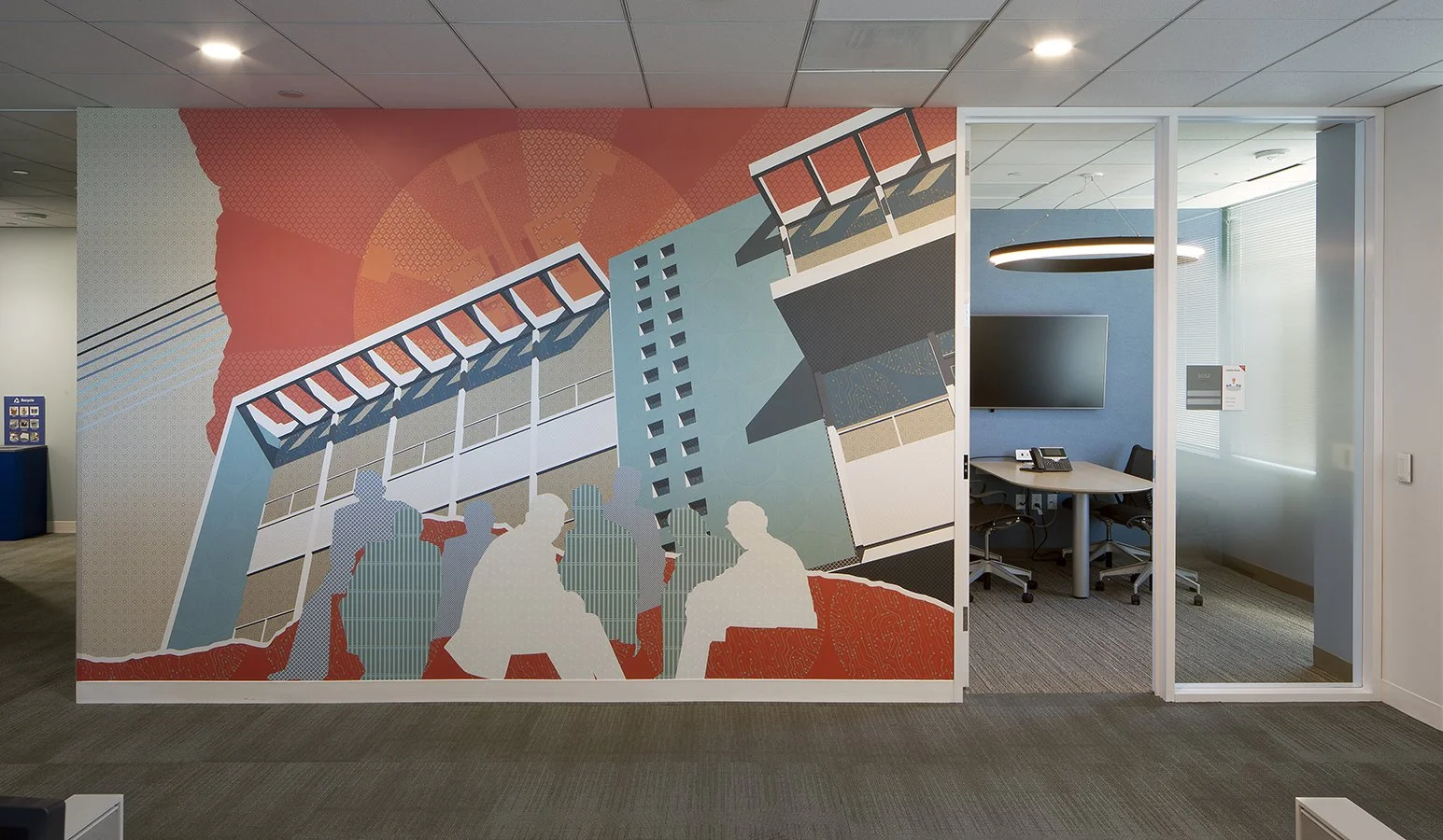 Office space with a colorful mural of people and buildings on a partition wall, and a meeting room with a round light fixture and a television in the background.