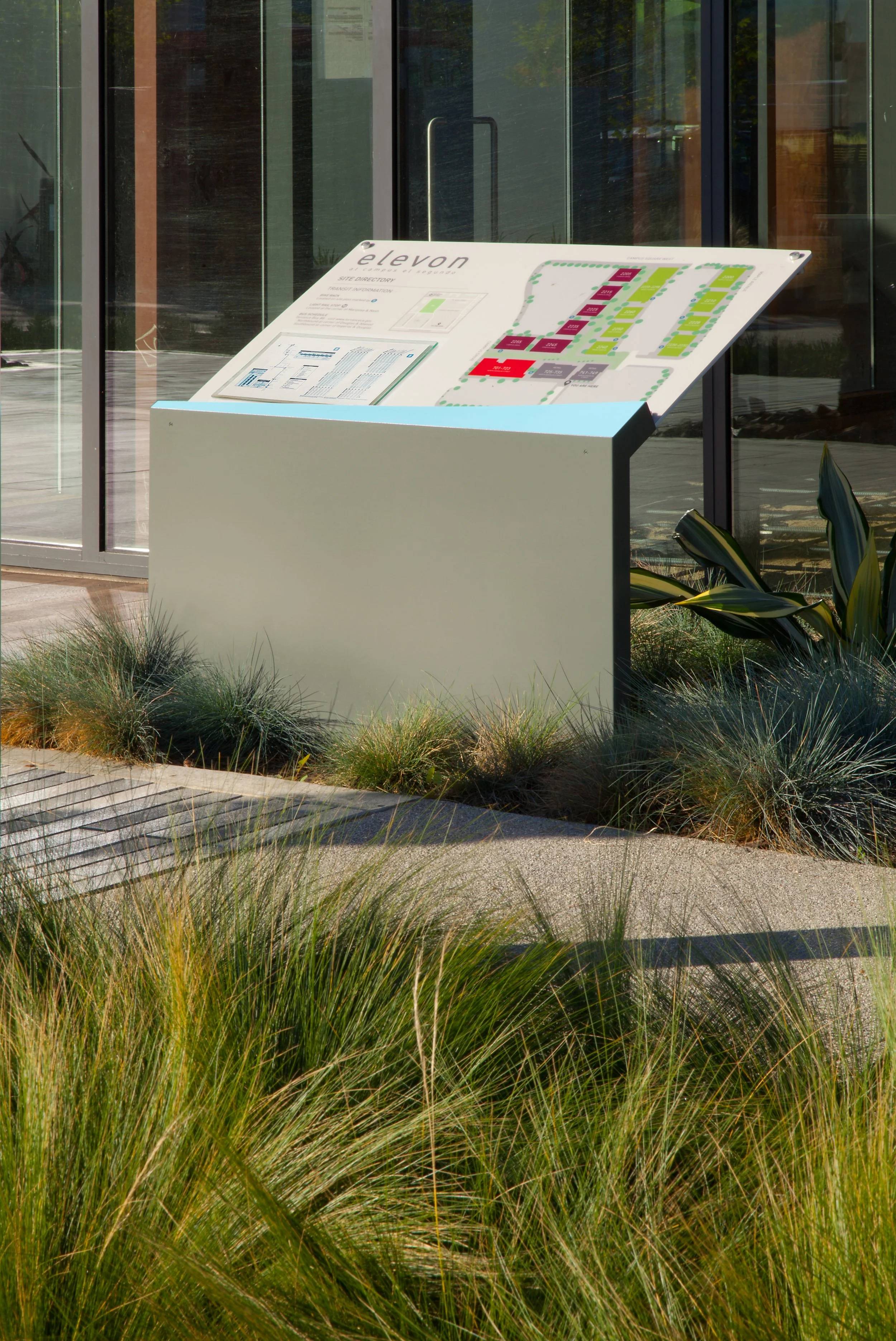 An outdoor informational kiosk with a map and directory, located in front of a glass building, surrounded by grass and plants.