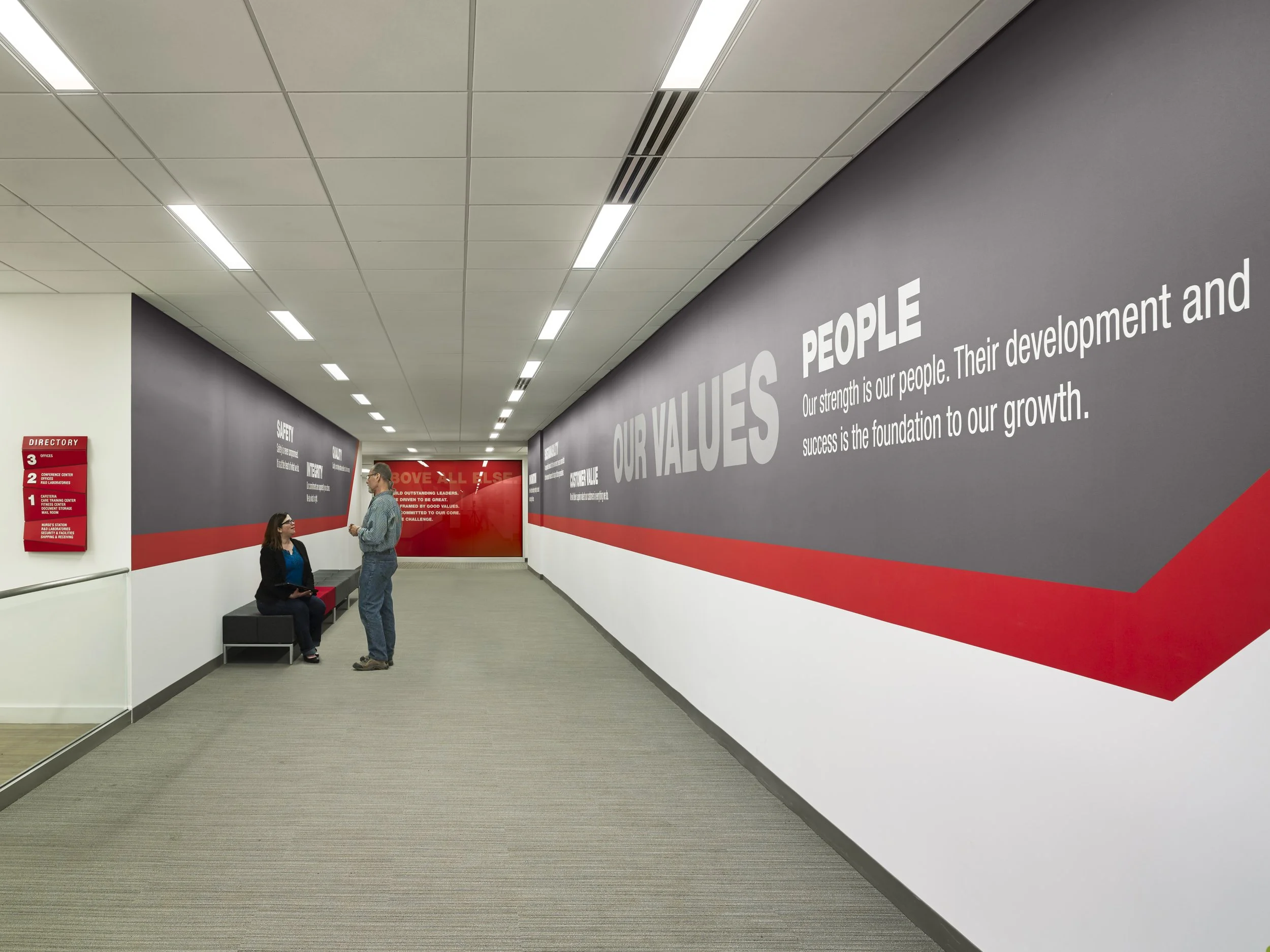 Interior hallway with white ceiling lights, purple and white walls with motivational words, a red directory sign, and two people talking near a bench.