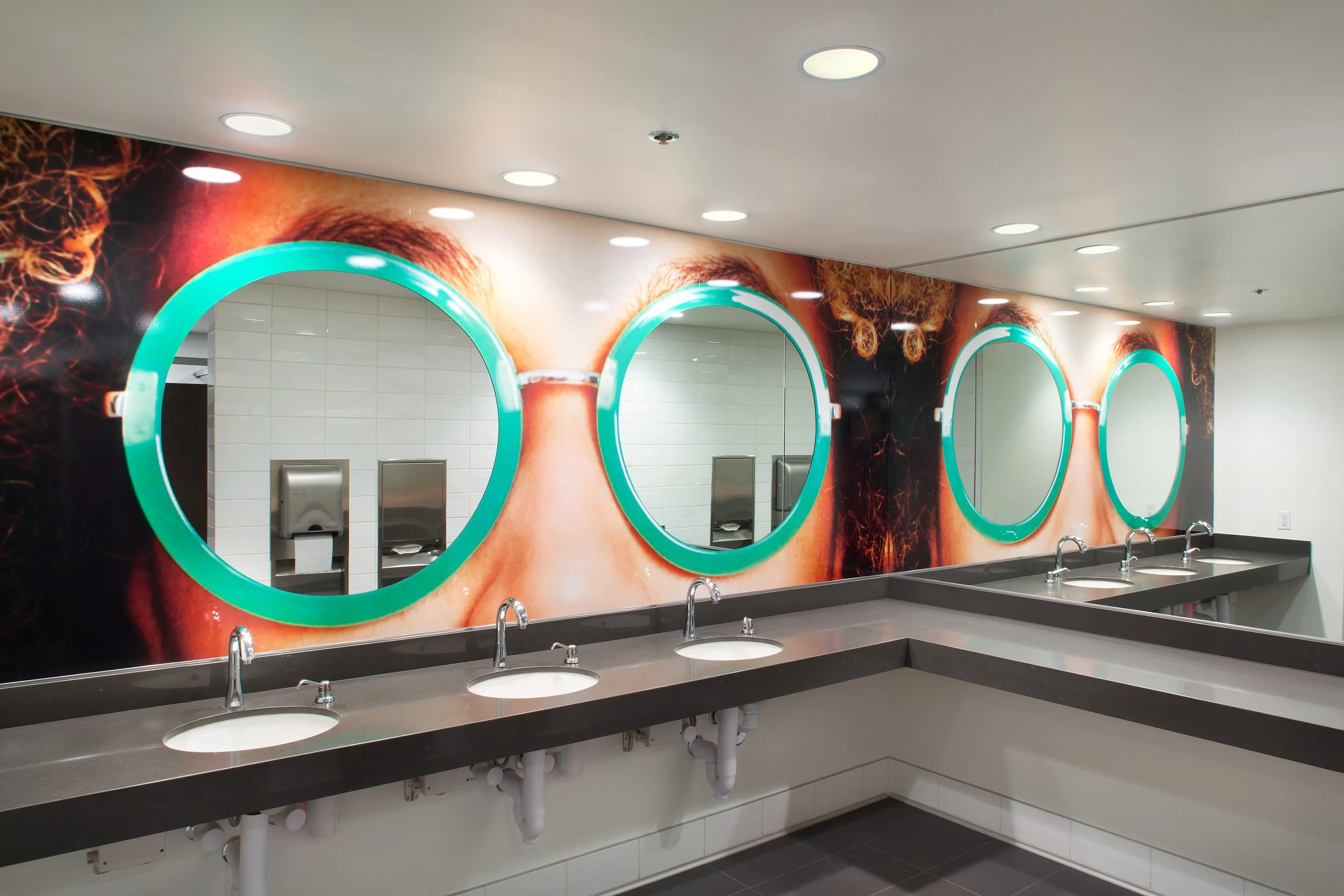 Public restroom with a large mural of a person's face wearing round teal neon-lit mirrors over individual sinks with faucets and soap dispensers.