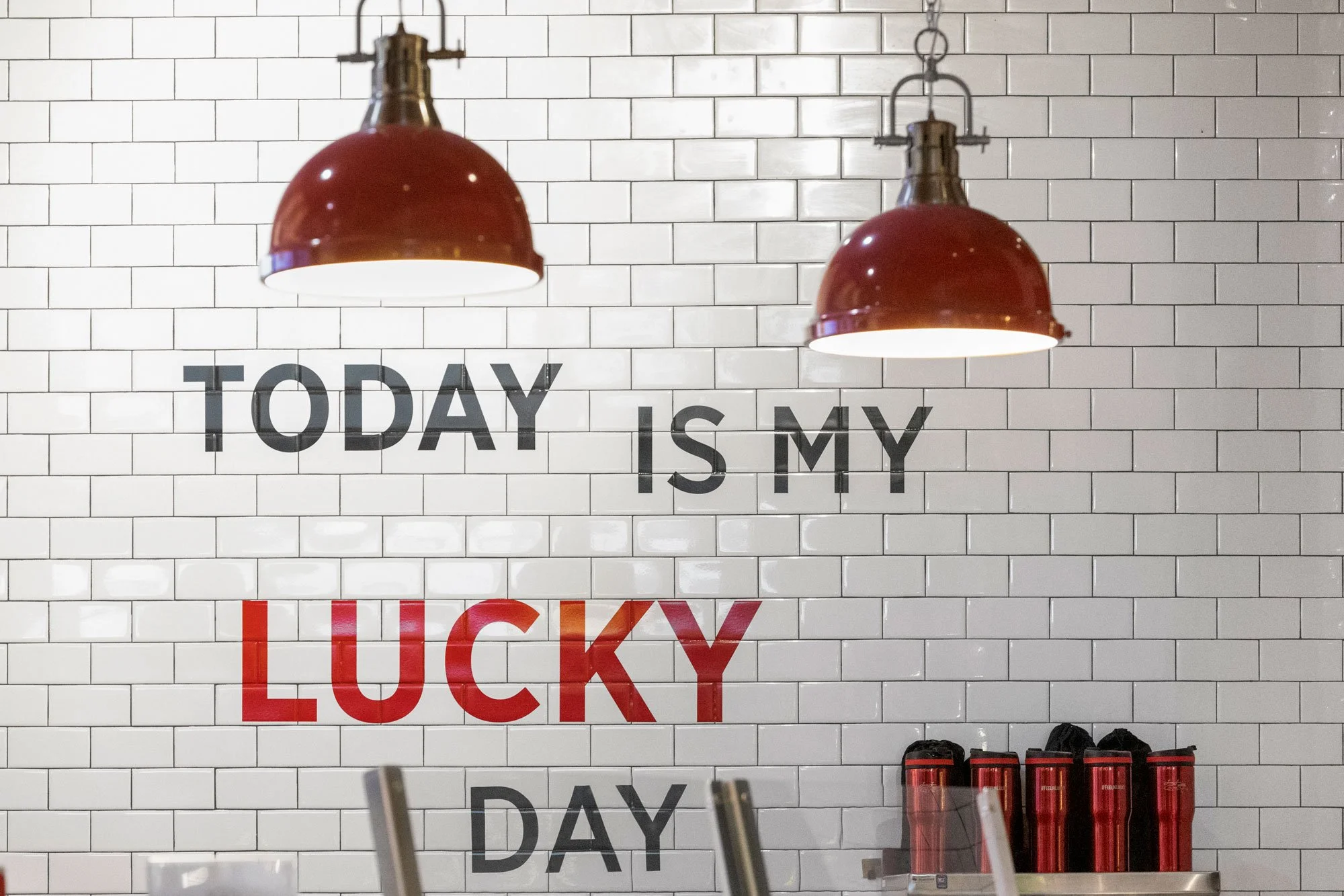 Two red pendant lights hanging above a white tiled wall with black letters spelling "TODAY IS MY LUCKY DAY" and red containers on a shelf below.