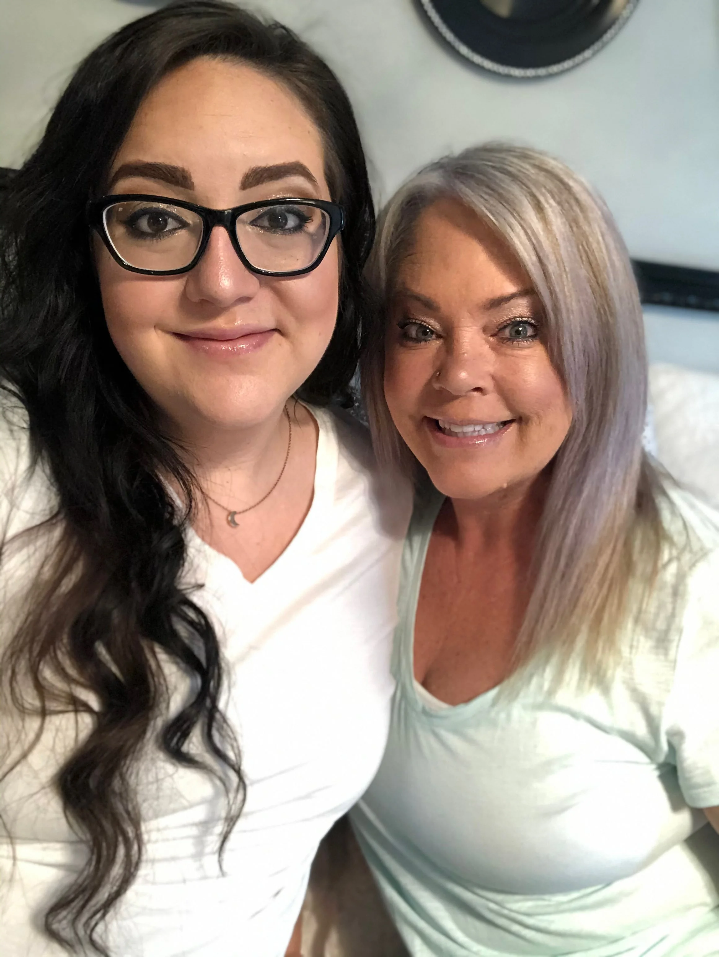 Close-up of two women smiling, one with black hair and glasses, and the other with gray hair, wearing light-colored tops, in an indoor setting.