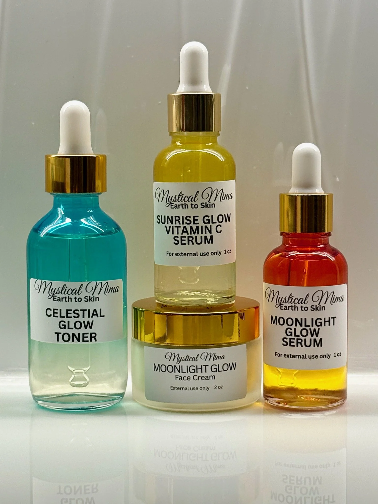Three skincare serum bottles with dropper caps placed on a reflective surface. The middle bottle is labeled 'Mystical Mima Earth to Skin Sunrise Glow Vitamin C Serum,' the left bottle is labeled 'Celestial Glow Toner,' and the right bottle is labeled 'Moonlight Glow Serum.'