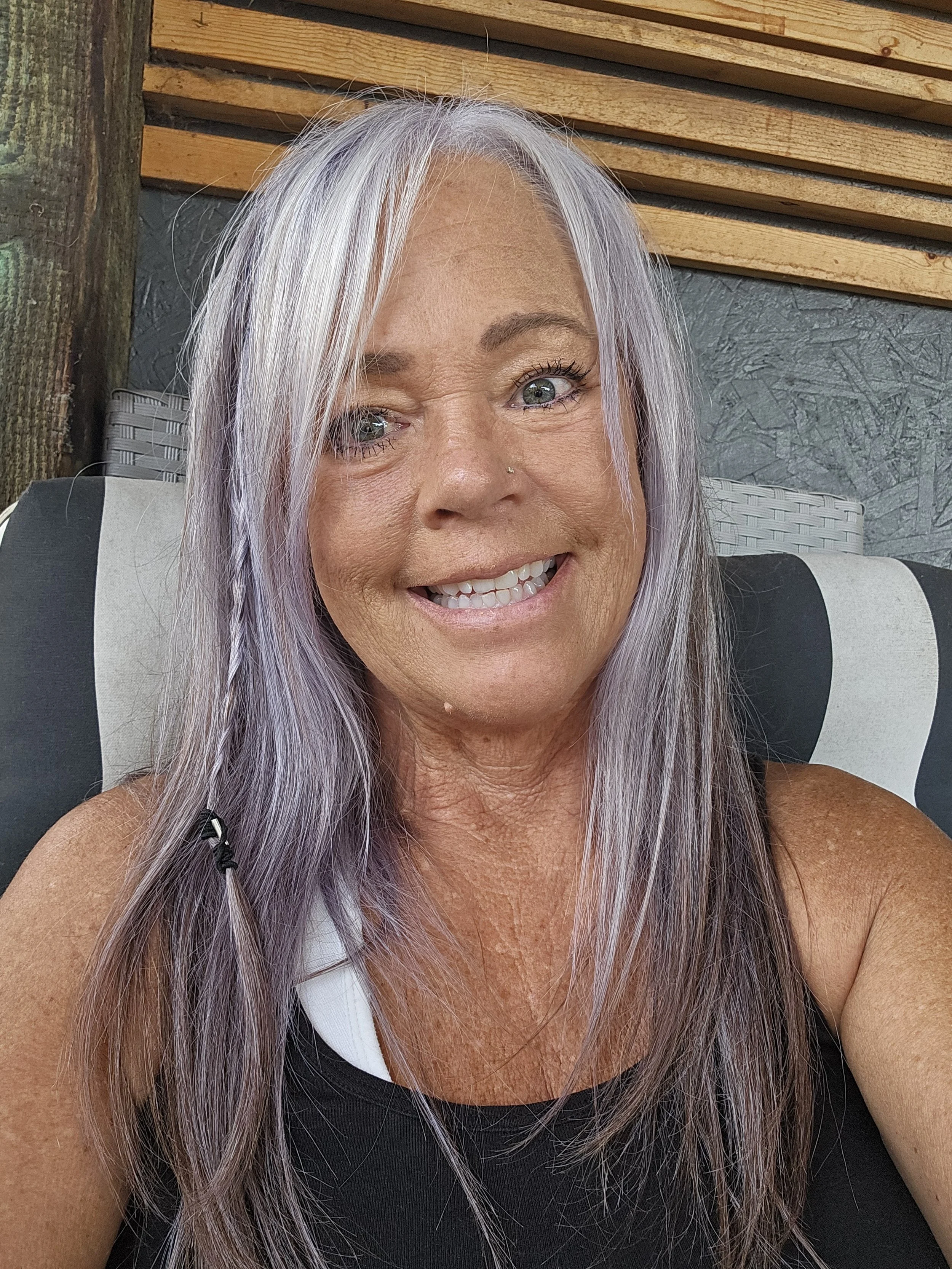 A smiling woman with long silver hair, wearing a black and white sleeveless top, sitting outside with a background of wooden and stone elements.