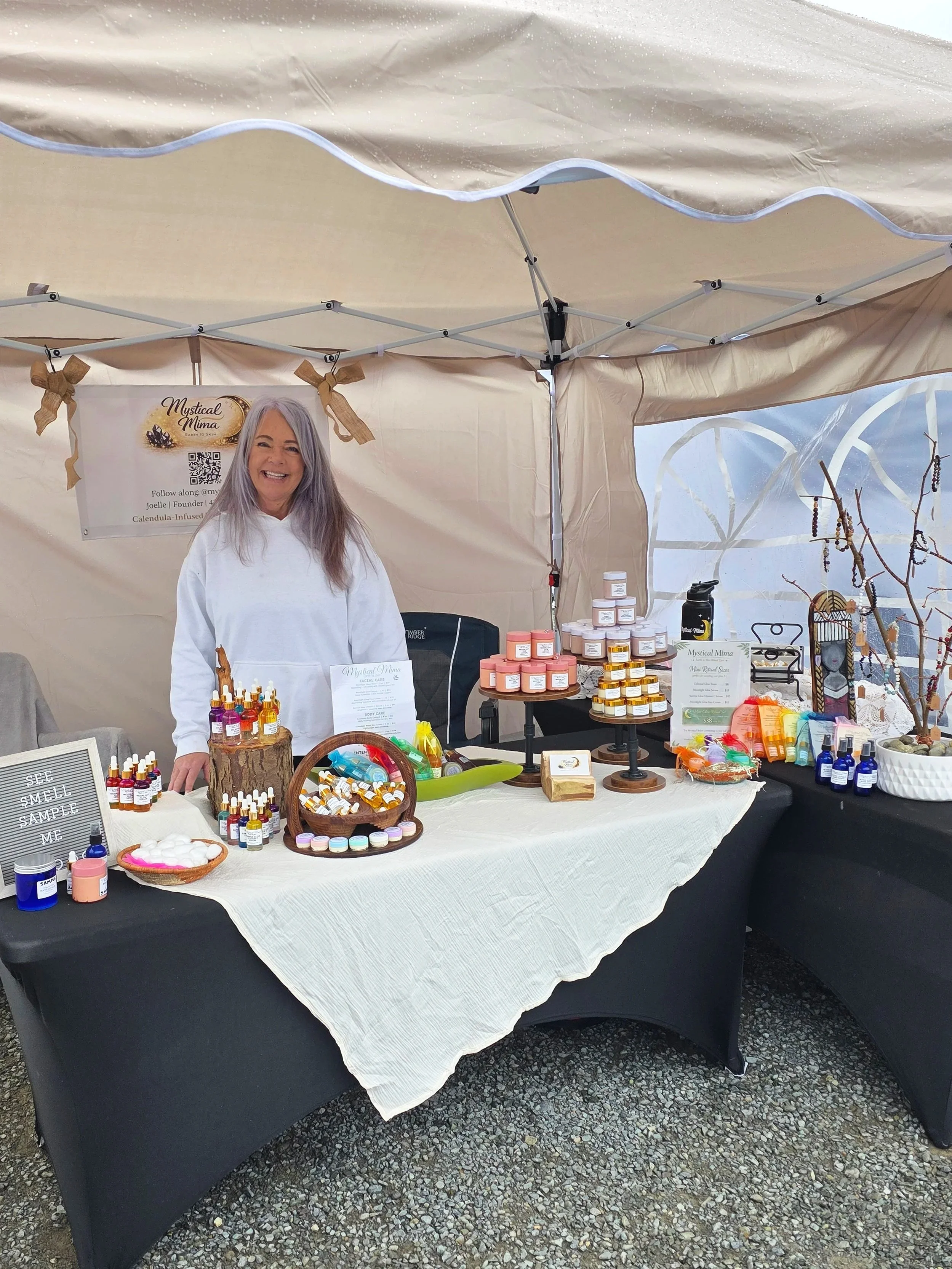 A woman standing behind a table at an outdoor market stall, selling skincare products. The stall has a variety of small bottles, jars, and containers, with a sign that reads "Mystical Mima" and a photo of a smiling woman with gray hair. There are decorative bows and a small potted plant on the table.
