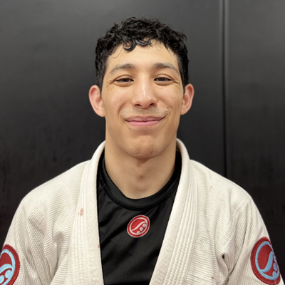 Young man smiling, wearing a jiu-jitsu gi with patches, standing against a dark background.