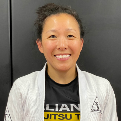 Grinning woman in a white martial arts gi and black T-shirt with yellow text, standing in front of a dark background.