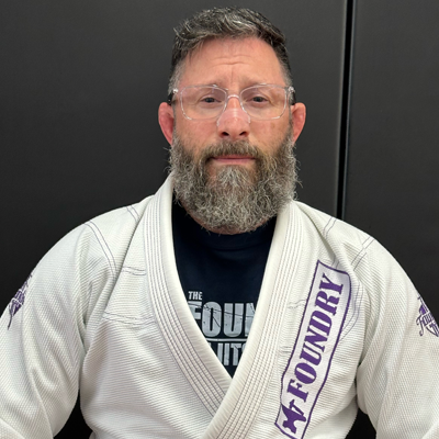 A man with glasses and a beard wearing a white martial arts gi with purple embroidery, standing against a black background.