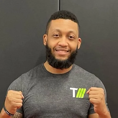 A man with a beard and short hair smiling, wearing a dark gray T-shirt and making fists in a fighting stance.