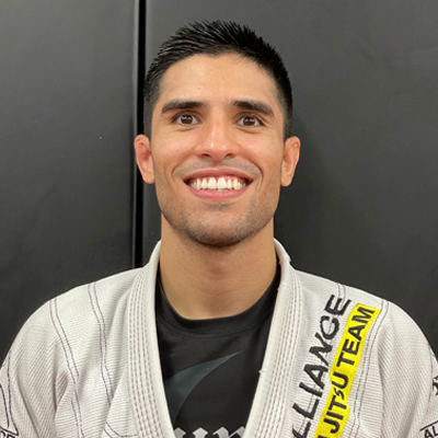 A young brazilian man in a white martial arts uniform with black and yellow patches, smiling and standing against a black background.