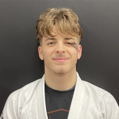 A young man with light brown hair and a slight smile, wearing a white jiu-jitsu gi over a black t-shirt, standing in front of a dark gray background.