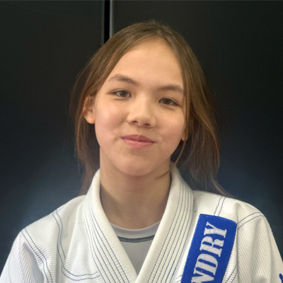 Young girl with light brown hair wearing a white martial arts gi with blue patches, standing against a dark background.