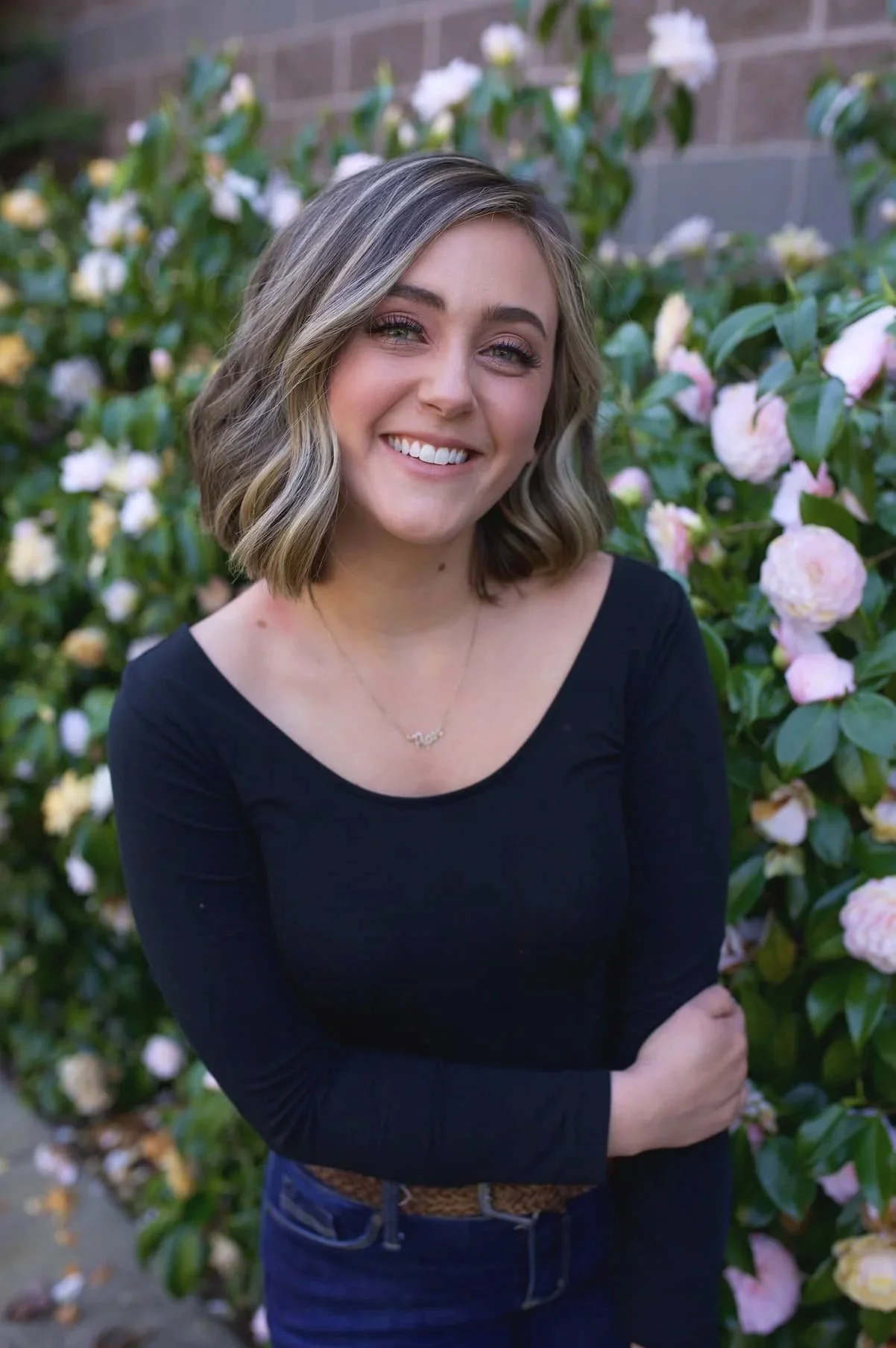 A young woman with shoulder-length wavy blonde hair smiling in front of a bush with pink and white flowers, wearing a black long-sleeve shirt and a necklace.