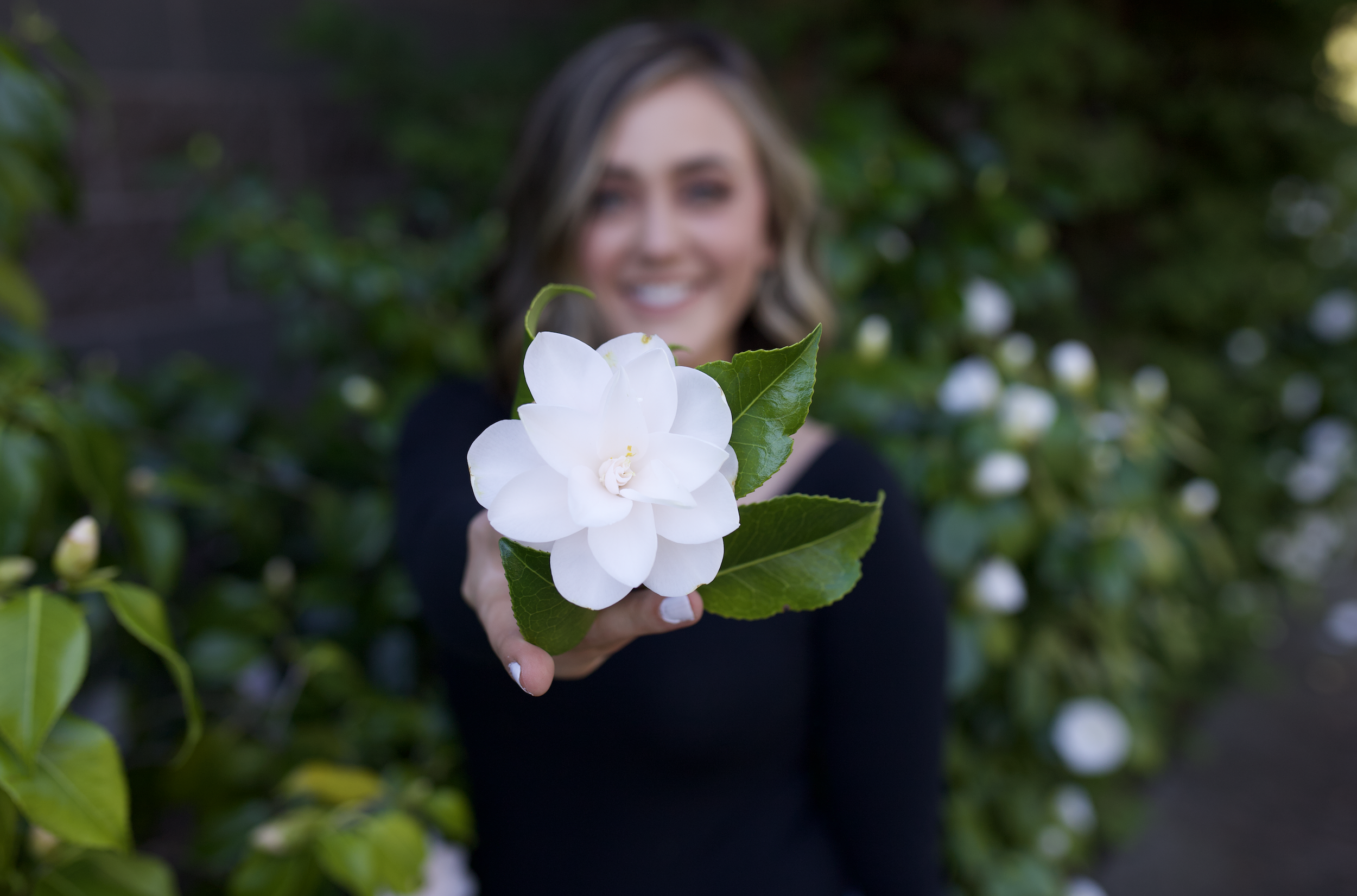 A woman with blonde hair and wearing a black top holds a white flower with green leaves in front of her, smiling in a garden with green foliage.