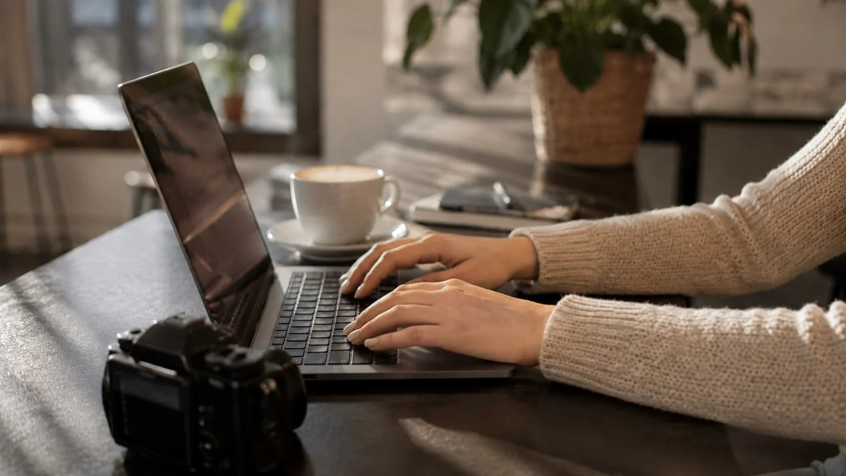 Person working on a laptop at a coffee shop table with a cup of coffee, a camera, and a plant in the background.