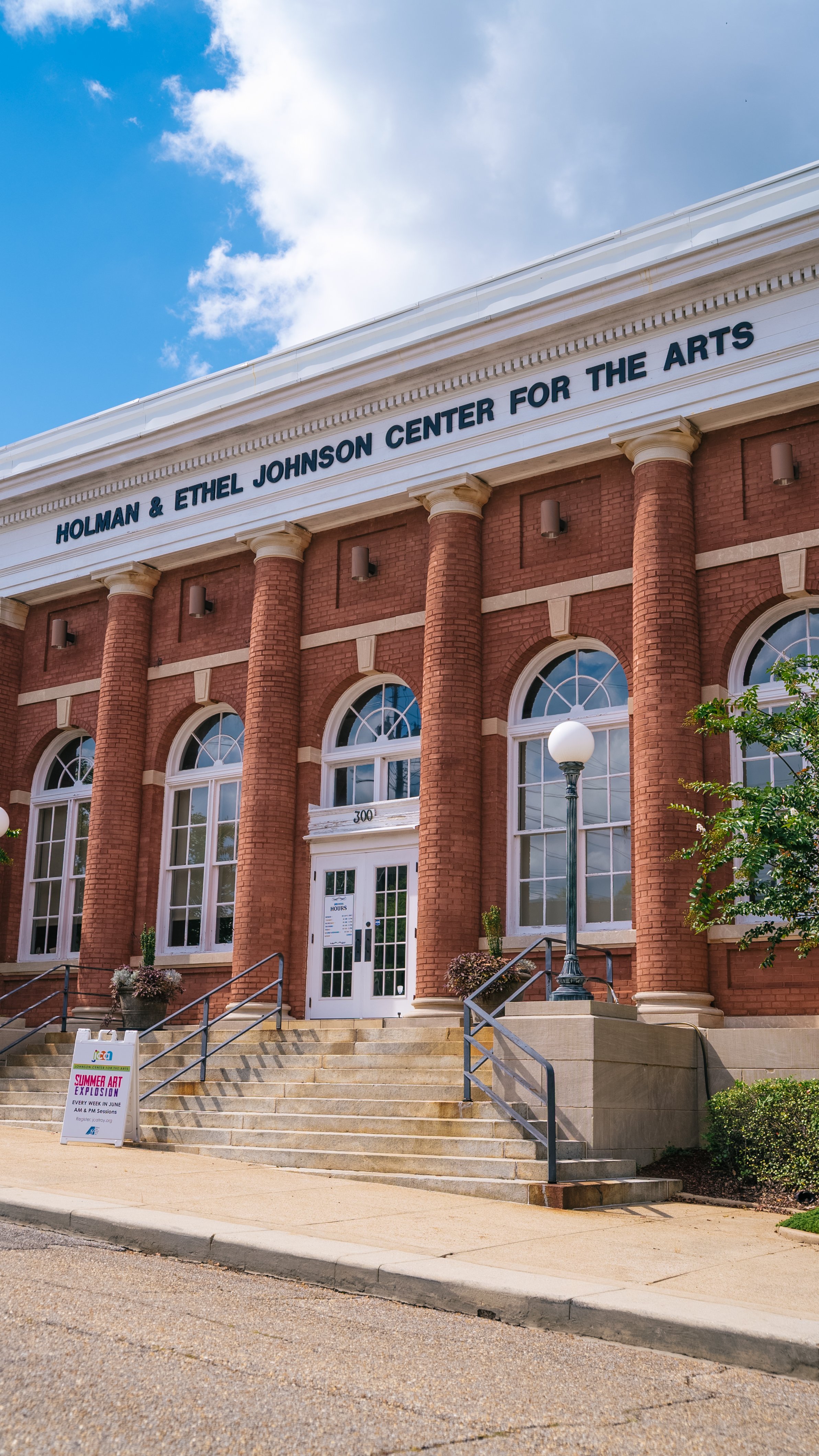 Front view of the Holman & Ethel Johnson Center for the Arts, a red brick building with large arched windows and white columns, steps leading up to the entrance, a streetlamp, and a sign for a summer art exhibition outside.
