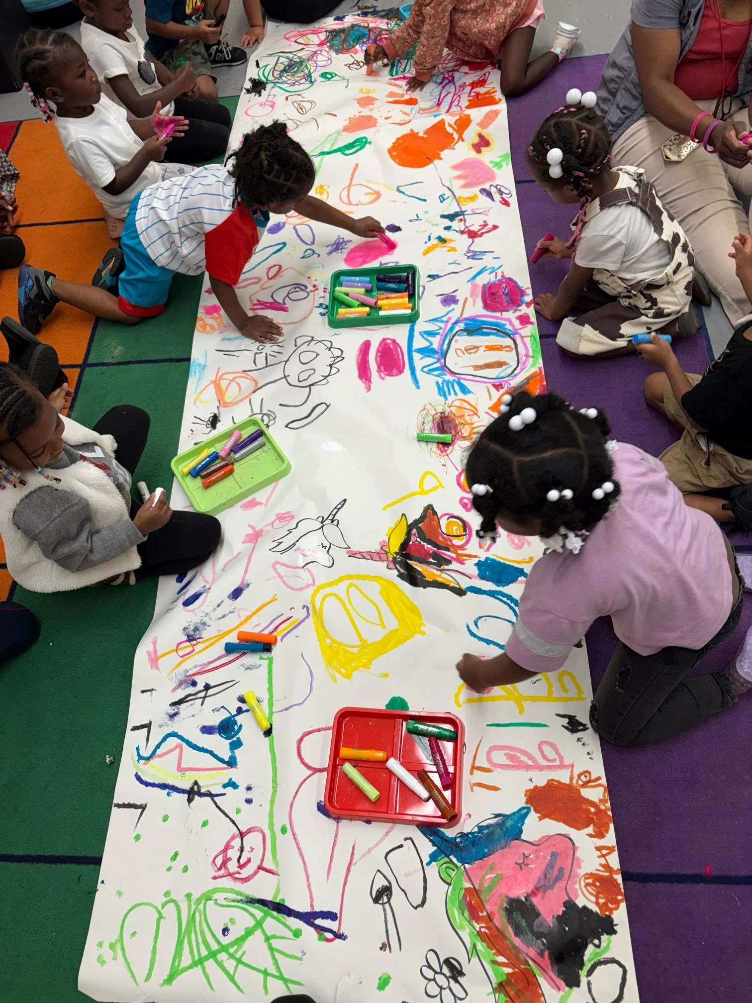 Children drawing and coloring on a large white paper with various colorful markers, some children kneeling or sitting on the floor, art supplies in containers, and a purple and orange floor mat.
