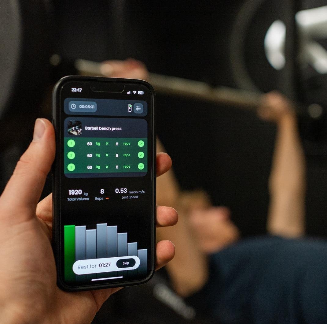 A person holding a smartphone displays a workout app during a gym session, focusing on a barbell bench press exercise, with workout details and a rest timer visible on the screen.