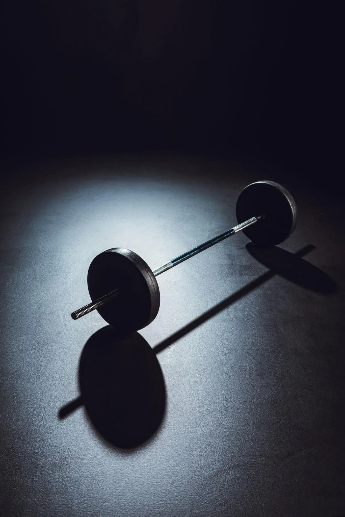 A dumbbell lying on a dark floor with a focused light source casting its shadow.