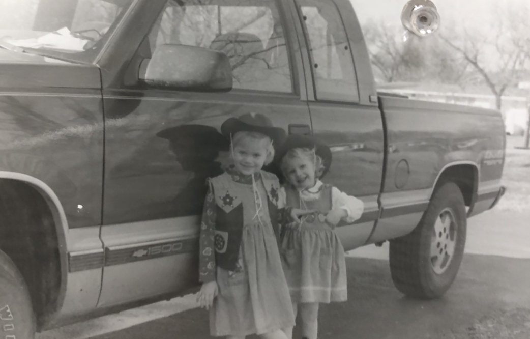 Two young girls dressed in costumes standing next to a pickup truck, with one girl holding a toy wand and smiling.