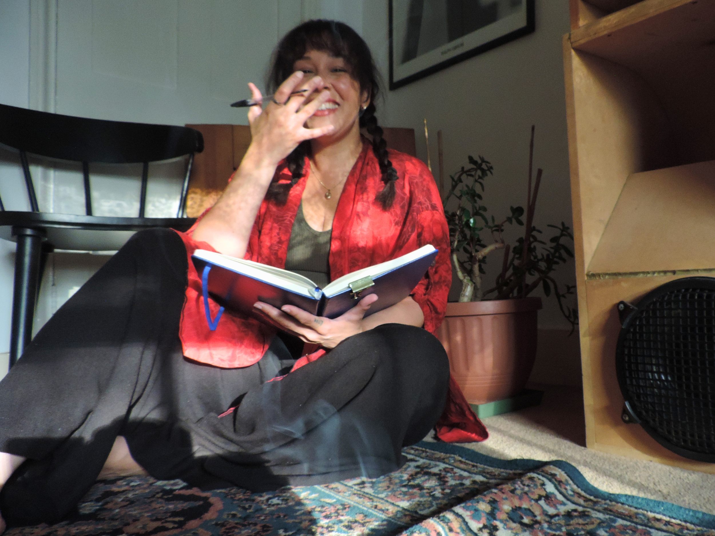 Woman with braids wearing a red jacket, sitting on a colorful rug, smiling while holding a notebook and a pen. Indoors, with plants and framed picture on the wall.