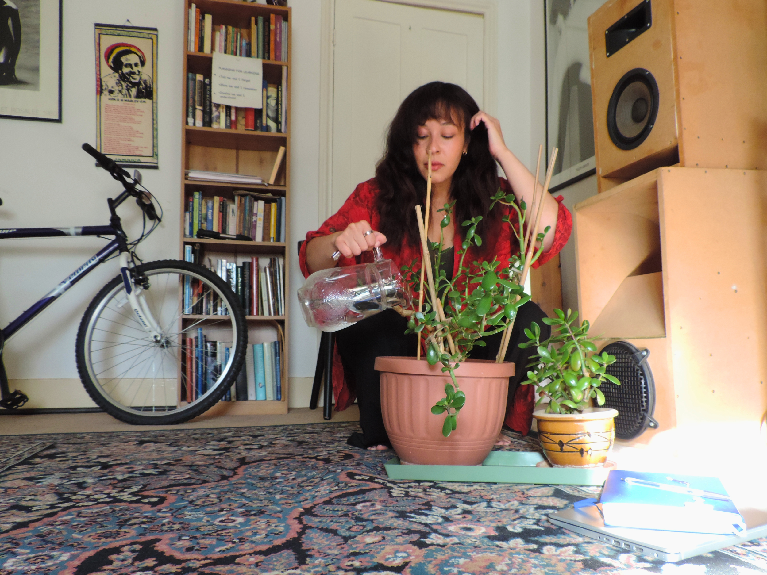 A woman with dark, wavy hair, wearing a red blouse, sitting on the floor tending to a large potted plant with green foliage. She appears to be watering or examining the plant. In the background, there is a white closed door, a bookshelf filled with books, and some posters on the wall. An orange speaker is seen on a wooden stand to the right, and a bicycle leans against the wall on the left. The scene is indoors, with a patterned rug covering the floor.