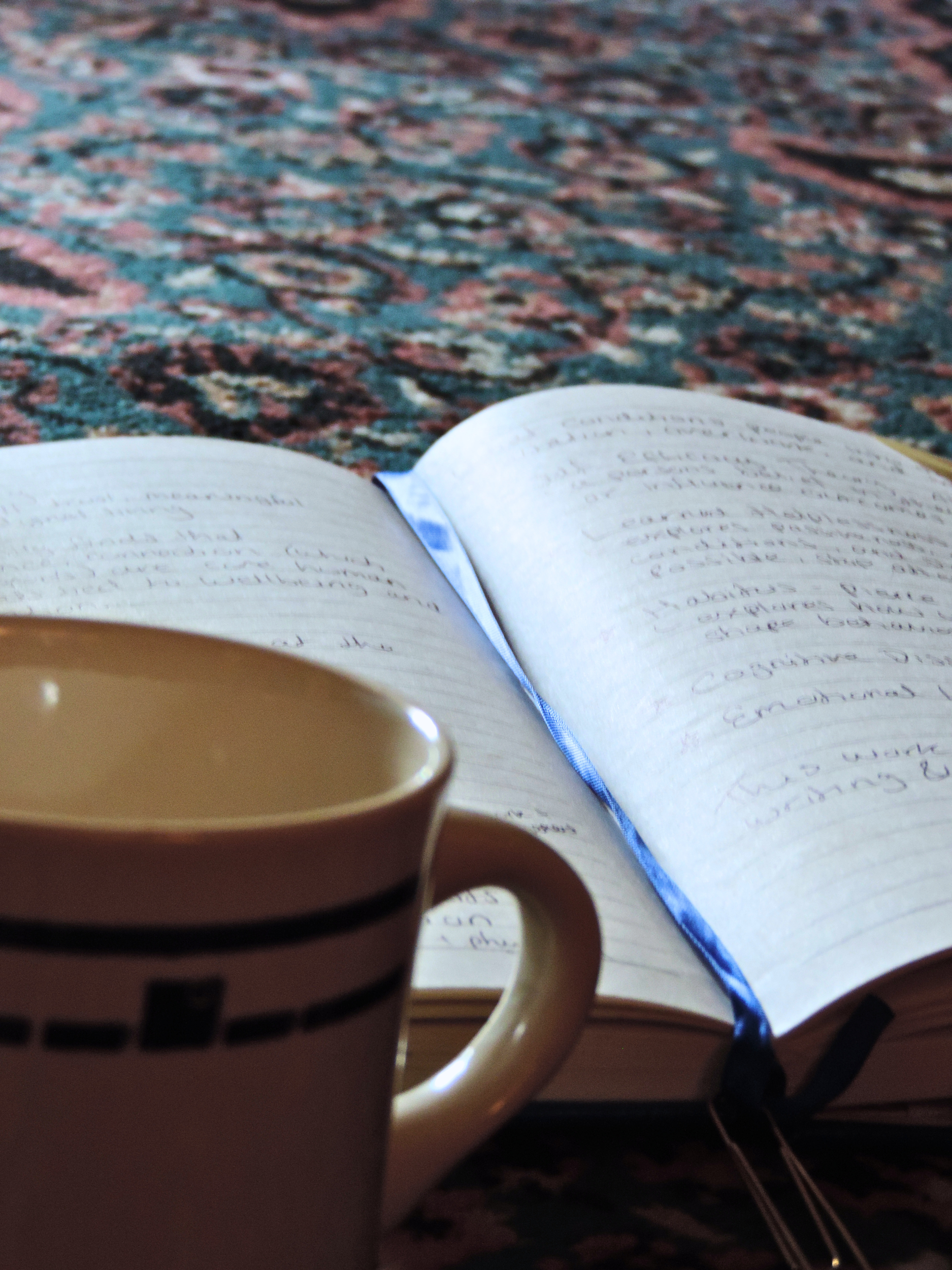Close-up of a white ceramic mug with a textured handle, partially covering an open notebook with handwritten notes, and a guitar neck in the background, on a patterned carpet.