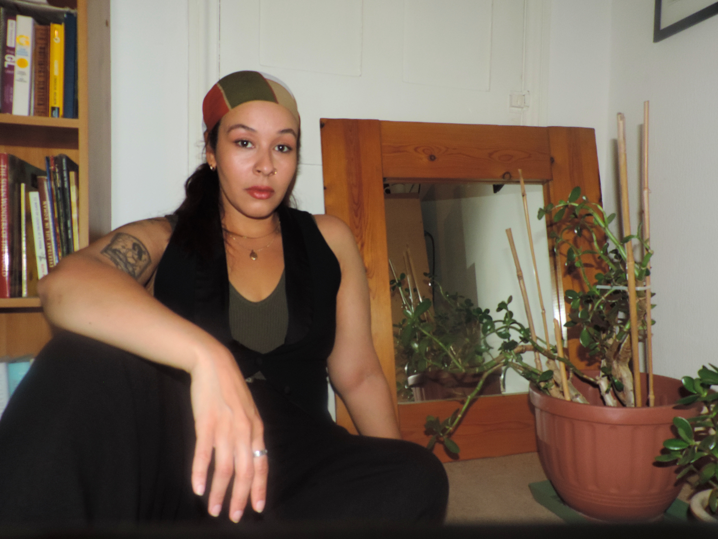 A woman with dark hair, wearing a multicolored headscarf, black sleeveless top, and gold jewelry, sitting indoors surrounded by potted plants and bookshelves.