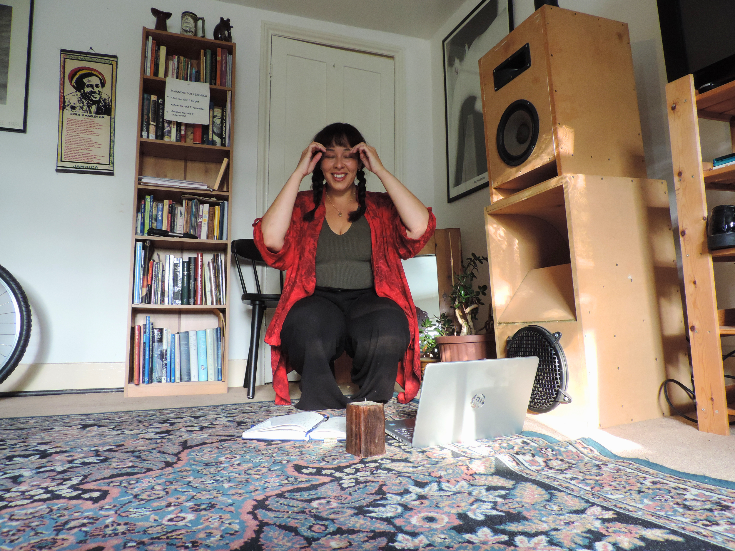 A woman with braids wearing a red shirt sitting on a black chair, smiling and touching her temples, in a room with bookshelves, artwork, a large speaker, a laptop, and a patterned rug.