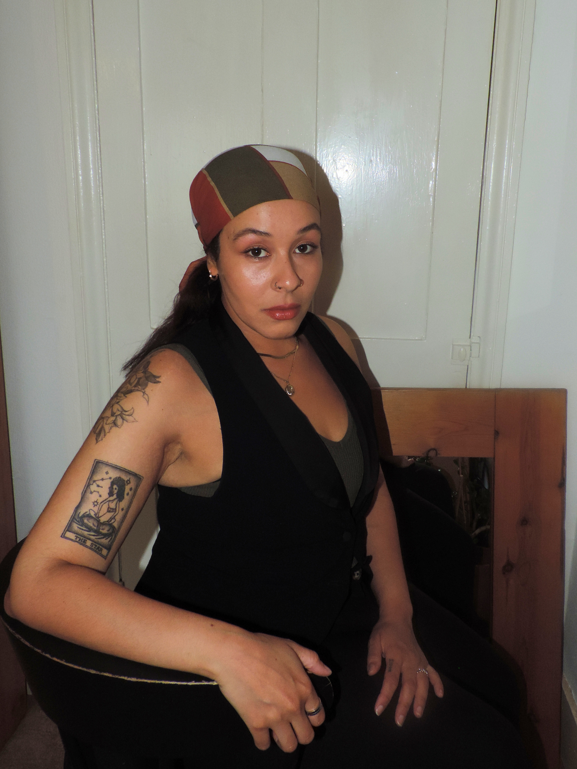A woman with tattoos on her arm wearing a patterned headscarf sitting in a room with white walls and wooden furniture.