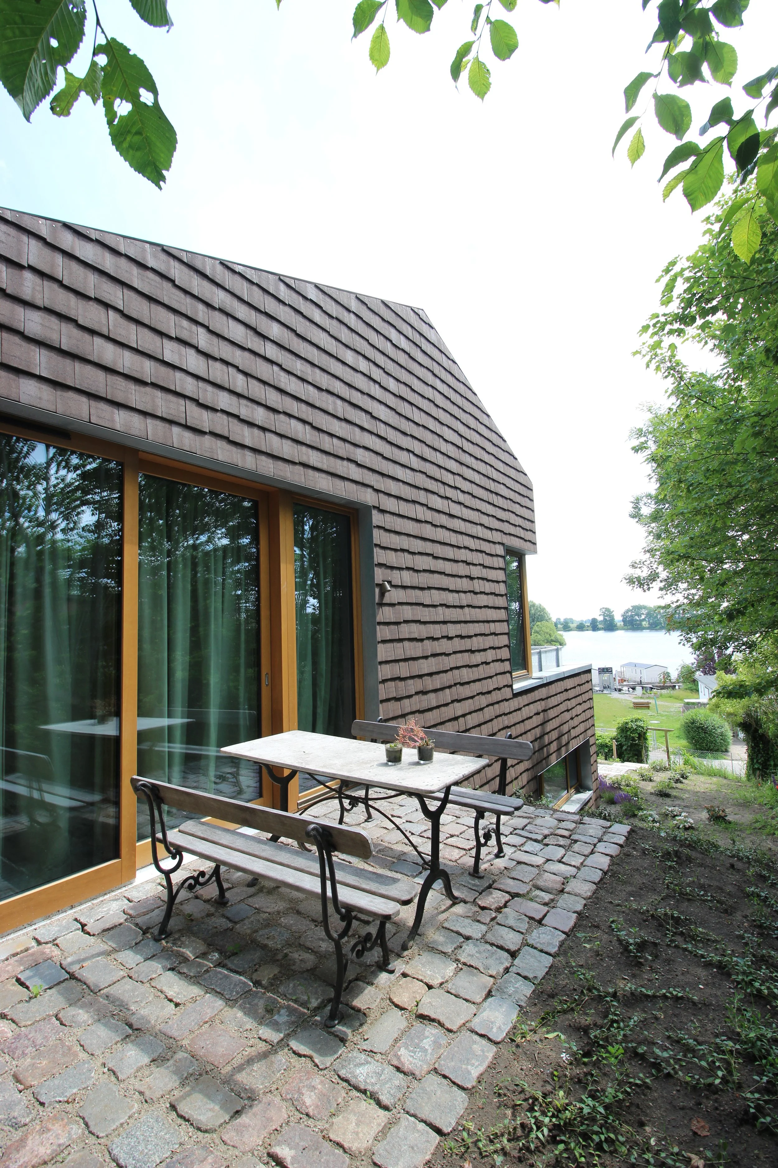 A patio with a stone tiled floor, a wooden table with potted plants, and benches. A house with brown shingle siding and large windows is visible. Trees and water can be seen in the background.