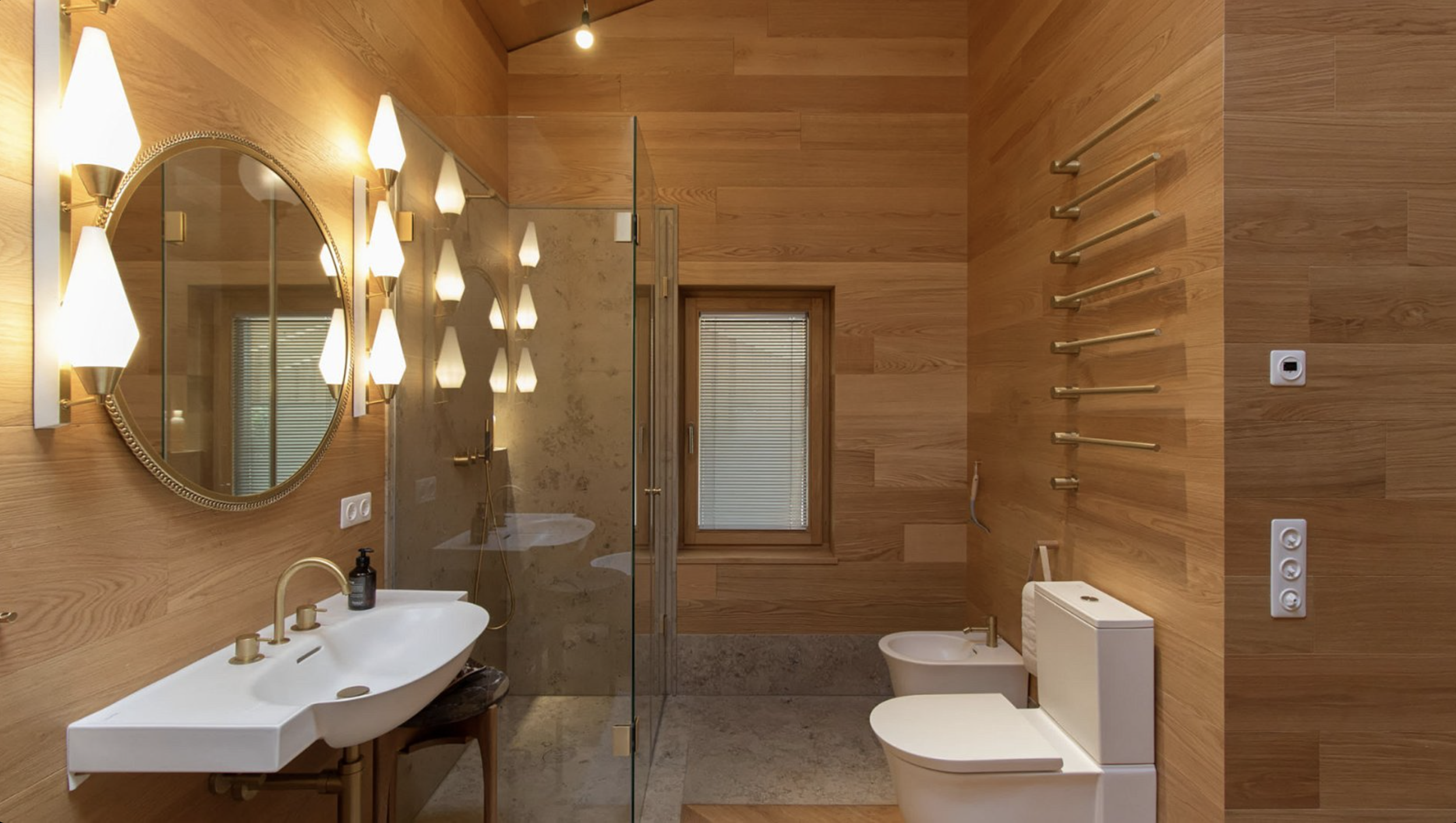 A modern bathroom with wooden wall panels, featuring a white vessel sink with brass fixtures, a large round mirror with decorative golden frame, and unique wall-mounted light fixtures. There is a glass shower enclosure behind the sink with beige marb
