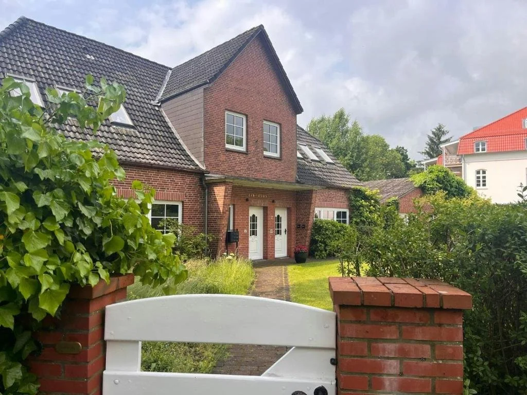 A brick house with a gray sloped roof and plants in front, viewed from behind a white gate and brick pillars.