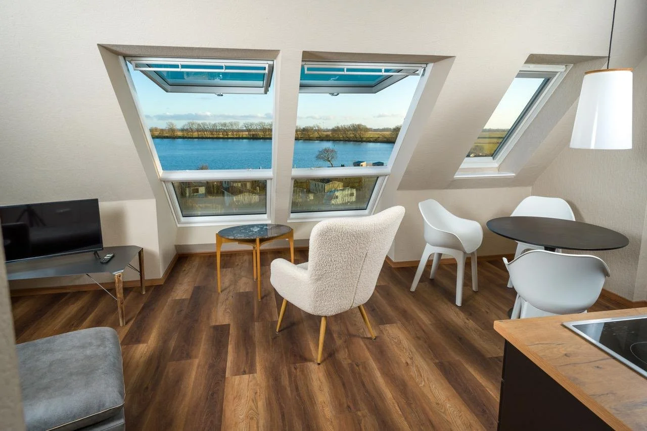 Room with three large skylight windows overlooking a lake, with a TV on a small stand, a white armchair, two white chairs at a black round table, and a hanging lamp.