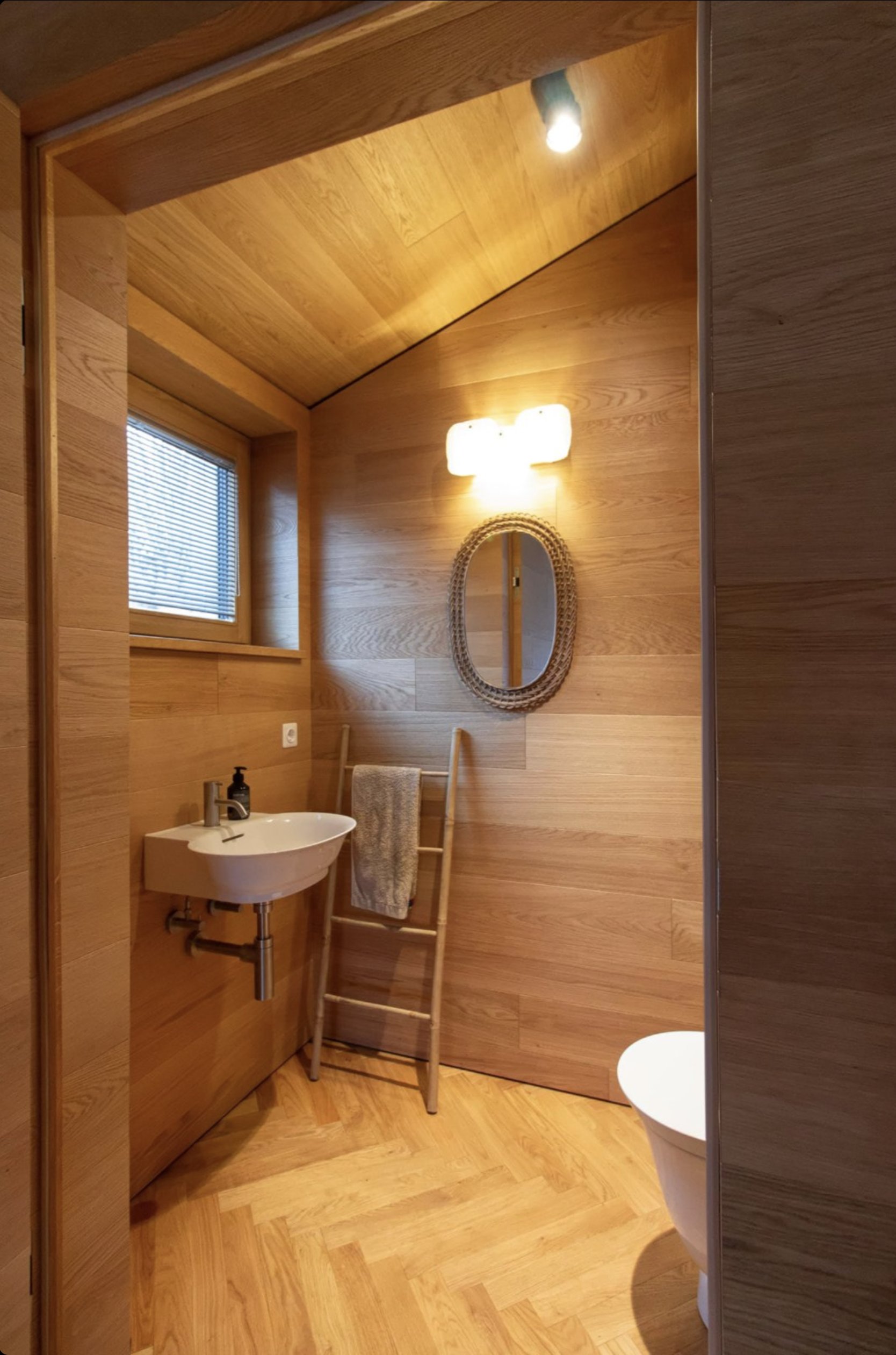 A cozy bathroom with wooden walls, ceiling, and floor. It features a small white sink with a black soap dispenser, a round mirror with a woven frame, a towel hanging on a ladder, a window with closed blinds, and a wall-mounted light fixture.