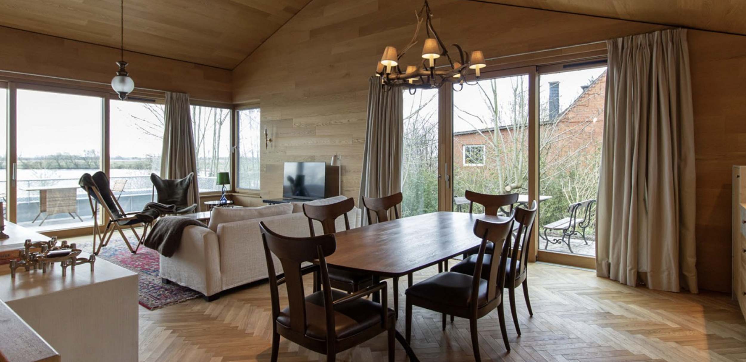 Living room with wooden ceiling, large windows, and sliding glass doors showing outdoor patio and trees. Contains dining table, chairs, sofa, armchair, TV, and chandelier.