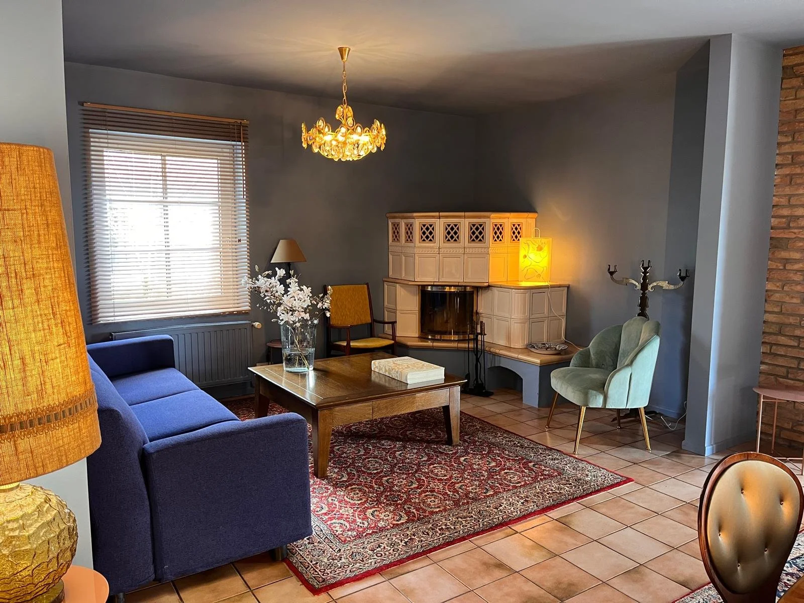 A cozy living room with a blue sofa, a wooden coffee table, yellow chairs, a fireplace, a chandelier, and various lamps, with a window and brick wall.