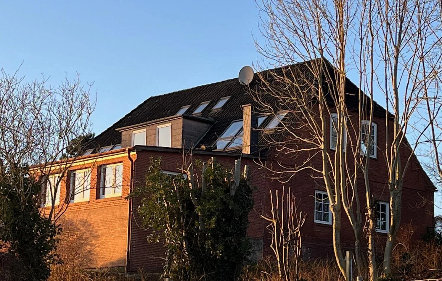 A multi-story brick house with a dark sloped roof, several skylights, and leafless trees in front, bathed in warm sunlight during dusk or dawn.
