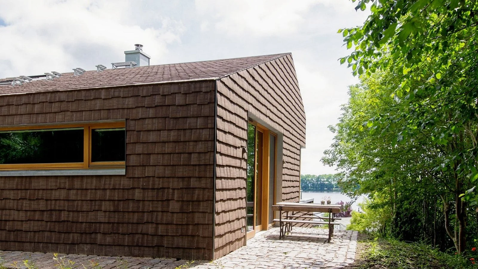 Modern house with brown tiled exterior wall, large window, glass door, and outdoor seating area on a cobblestone patio surrounded by lush green trees, overlooking a body of water.