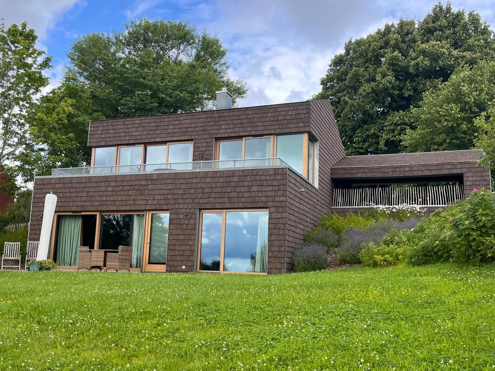 Modern brown house with large glass windows and a balcony surrounded by greenery and trees under a partly cloudy sky.