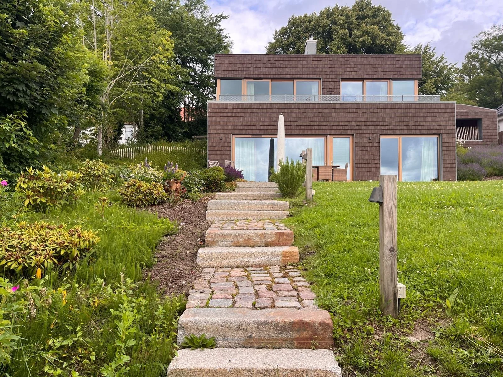 Brick house with large glass sliding doors and windows, surrounded by a green garden with plants and flowers, a stone staircase leading up to the house, and a pathway with cobblestones in the front yard.