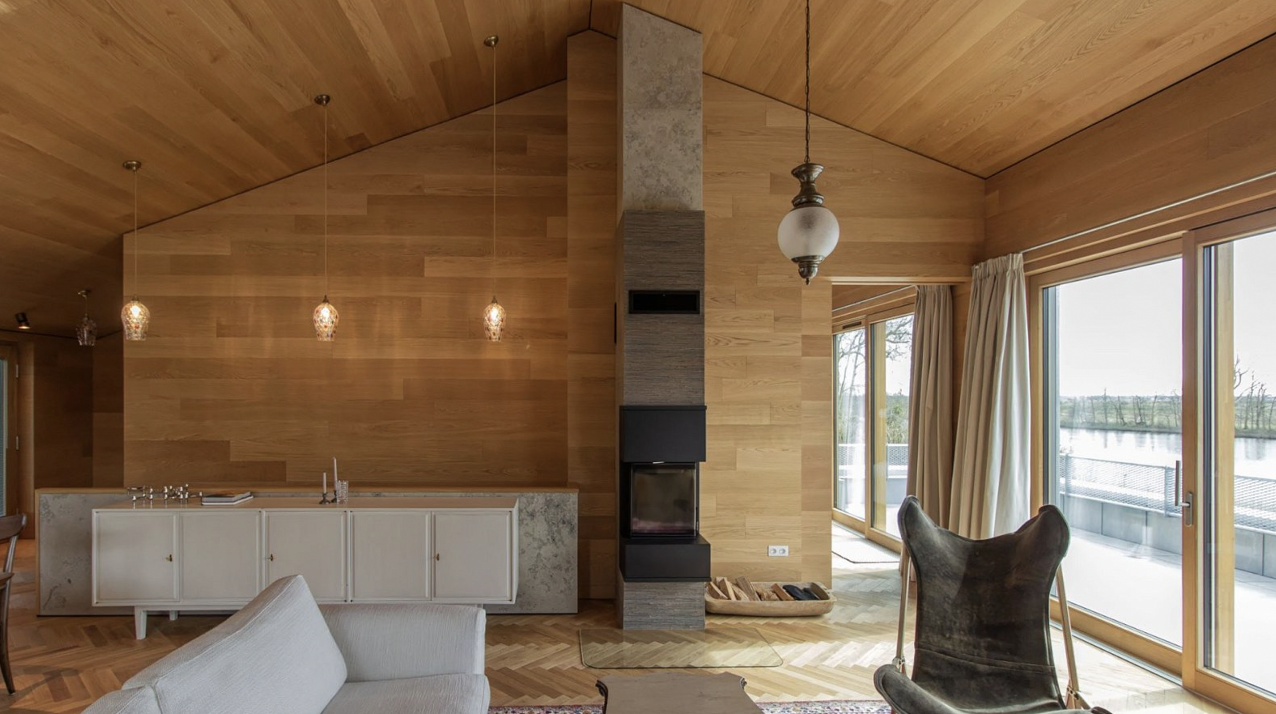 Interior view of a modern living room with wooden walls and ceilings, large glass doors with curtains, and a view of a water body outside.