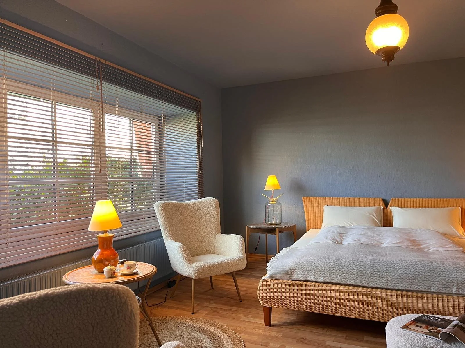 Cozy bedroom with large window, wooden blinds, beige armchair, wicker bed with white bedding, wooden nightstand with small lamp, other small furniture, and warm lighting in an inviting space.