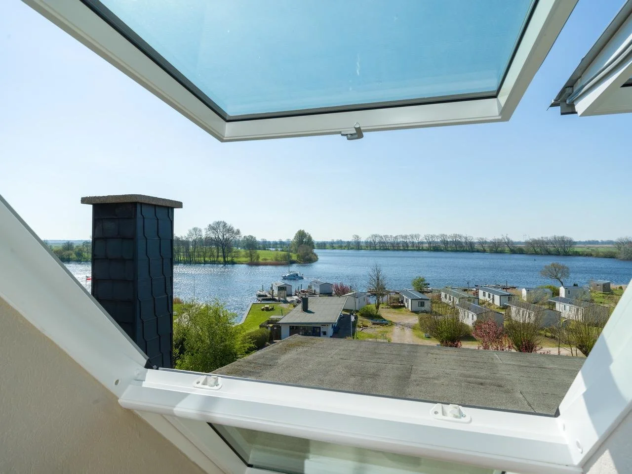 View from an open attic window overlooking a river with houses, trees, and a boat in the distance on a sunny day.