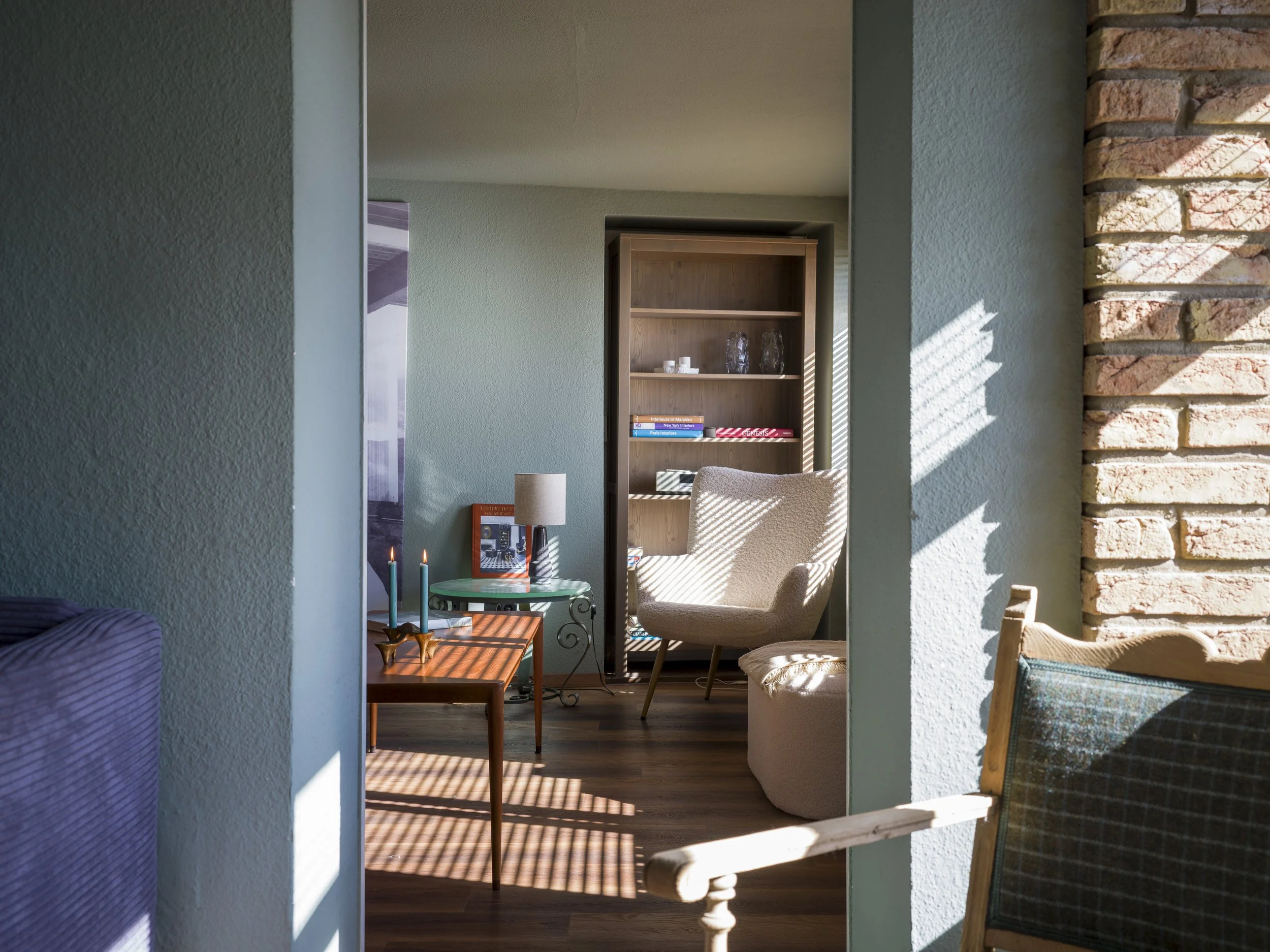 A cozy living room viewed through an opening, featuring a beige textured armchair, a small tip-over table with candles, and a wooden bookshelf with decor items. Sunlight casts shadows through blinds onto the furniture and wooden floor.