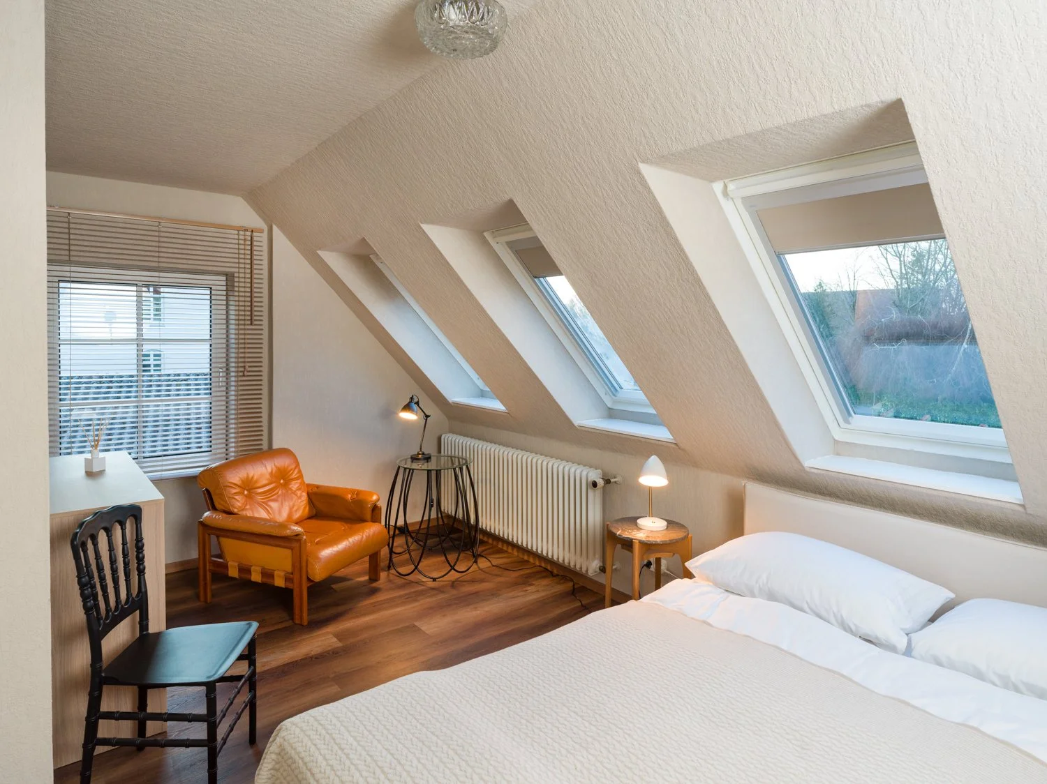 A bedroom with slanted ceilings and three skylight windows. The bed has white bedding, and there are two small lamps on tables beside it. There are two chairs, one black and one tan, near a window with blinds, and a small white desk.