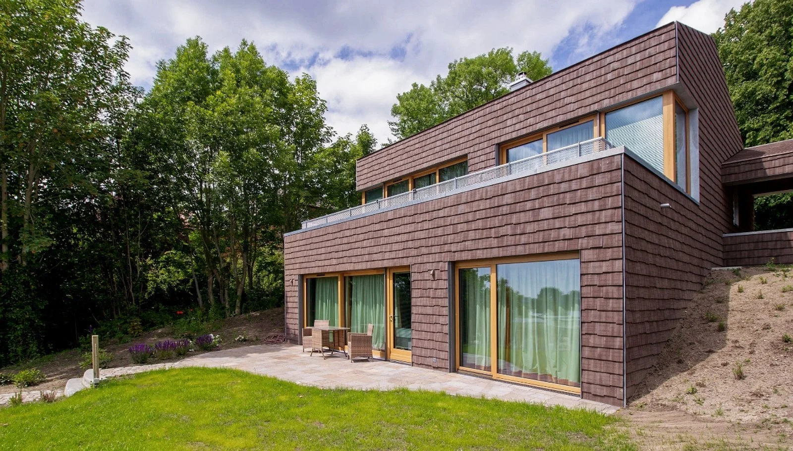 Modern multi-level house with brown shingle exterior, large sliding glass doors, balcony, and outdoor patio furniture, surrounded by green trees and lawn.